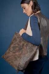 Bolso tipo shopper de cuero color suela con textura grabada, detalle de costuras laterales trenzadas y placa metálica con logo de la marca. Cuenta con doble asa larga para colgar al hombro, cierre principal y múltiples bolsillos internos y externos.