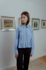 Camisa celeste de poplin de algodón, corte irregular con tablas al frente, cuello clásico y mangas largas.