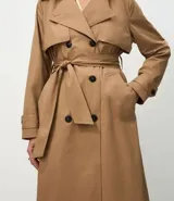 Trench coat largo de Marfinno, confeccionado en gabardina de poliéster. Presenta cuello con solapas, aletas en los hombros, cierre frontal con botones y cinta para lazo en la cintura.