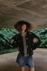 Camisa negra de manga larga con cuello redondo y escote en V, adornada con bordados florales en contraste en tonos beige y marrón a lo largo de la abertura frontal, hombros y mangas.