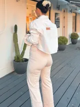 Camisa blanca de algodón con corte cropped y calce relajado. Presenta cuello clásico, mangas largas con puños anchos y una serie de parches bordados estilo militar e insignias metálicas decorativas en el frente y las mangas.