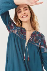 Vestido amplio color azul petróleo con bordados étnicos en tonos rojo, blanco y azul oscuro en el canesú y mangas. Presenta escote en V con cordones de ajuste terminados en borlas rojas y doradas.