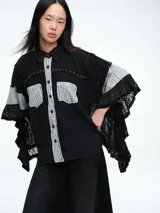 Camisa negra de corte holgado con mangas tipo murciélago, detalles de encaje en los bordes y paneles con estampado floral gris en la botonera y bolsillos frontales. Incluye apliques de tachas metálicas en la parte superior del pecho.