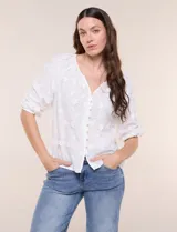 Blusa blanca de calce holgado, con bordado en relieve tipo broderie de motivos florales en toda la prenda. Presenta escote en V con fruncido en los hombros, cierre frontal con botonadura y mangas tres cuartos abullonadas con puños elastizados.