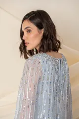 Vestido largo de tul en color celeste pastel, con escote en V y breteles anchos. Presenta un diseño bordado con lentejuelas y pedrería en líneas verticales y una capa larga integrada que nace desde los hombros.