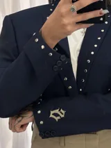 Blazer corto de color azul marino, con solapas decoradas con tachas metálicas plateadas y detalle de dije metálico en la manga. Presenta un corte estructurado y cierre frontal.