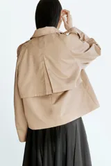 Gabardina corta color beige, de estilo cruzado con doble botonadura, solapas de muesca, bolsillos ribeteados y mangas raglán con charreteras en los hombros.