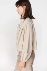 Blusa de algodón color beige con cuello camisero, mangas largas abullonadas con puños elásticos y cierre frontal mediante tres lazos para anudar.