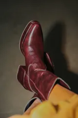 Bota estilo tejano de cuero color bordó con acabado brillante, taco cuadrado y punta cuadrada. Presenta costuras decorativas en el empeine y caña media.
