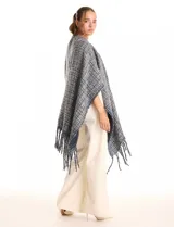 Ruana de tejido tipo tweed en tonos azul y gris con destellos de lurex. Presenta un diseño abierto con terminaciones desmechadas y flecos en todo el borde inferior.