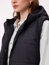 Chaleco largo acolchado color beige, con capucha forrada en sherpa, cierre frontal con cremallera y botones a presión, y bolsillos laterales.