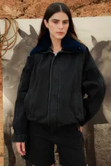 Campera oversize de jean color negro con cuello de corderito azul, cierre frontal con cremallera y solapa con botones a presión. Presenta mangas amplias y un pequeño bordado rojo en el puño.