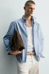 Camisa de hombre de manga larga, corte relajado (relaxed fit), color celeste con patrón de cuadros pequeños en azul y rojo. Presenta cuello abotonado (button-down), bolsillo de parche en el pecho y cierre frontal de botones.