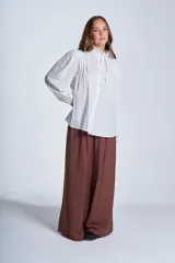 Camisa de color azul con cuello alto decorado con volados y lazo de ajuste. Presenta tablas decorativas en los hombros, cierre frontal con botones de nácar y mangas largas con puños elásticos que generan un efecto abullonado.