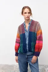 Chaqueta de tejido de punto con diseño multicolor a rayas diagonales en tonos cálidos y fríos. Presenta un panel frontal central en color azul sólido con flecos en el ruedo y puños tejidos en color violeta.