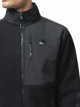 Campera de polar negro con cuello alto y cierre frontal completo. Presenta paneles de nylon en contraste en el pecho, hombros y codos, incluyendo un bolsillo vertical con cierre en el pecho y dos bolsillos laterales abiertos.
