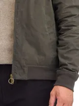 Campera de algodón encerado en color verde oliva, con cierre frontal, puños y ruedo elásticos, y cuello de pana en color marrón con detalle de broches metálicos.