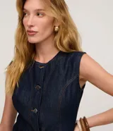 Chaleco largo de jean azul oscuro, con corte acinturado, cierre frontal abotonado y costuras con pespuntes en contraste. Presenta un diseño sin mangas con cuello redondo.