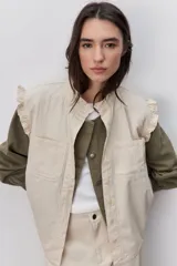 Chaleco de algodón color beige con calce relajado, cuello redondo y cierre frontal con botones. Presenta volados en los hombros y bolsillos delanteros amplios.