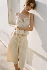 Top de color crema con escote en pico, tirantes anchos y cierre frontal de botones. Presenta bordados florales en tono verde claro alrededor del escote y en forma de semicírculo en la parte inferior delantera.