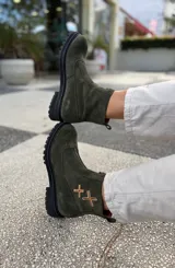 Botas cortas de cuero gamuzado color marrón oscuro, con suela track de goma negra y cierre frontal. Presentan un detalle de dos cruces bordadas a mano en tonos naranja y rojo en el lateral exterior.