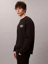 Buzo Calvin Klein de felpa color negro, con cuello redondo, manga larga, puños y dobladillo acanalados. Presenta un bordado frontal con la palabra "VERY" en tonos verdosos y debajo el logo de la marca.