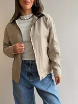 Campera de gabardina color beige con cierre frontal y cuello en contraste color negro. Presenta un diseño minimalista con bolsillos delanteros funcionales y calce cómodo.