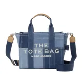 Bolso tipo tote de Marc Jacobs, modelo Mini Traveler, confeccionado en denim con diseño de patchwork en diferentes tonos de azul. Presenta el logo de la marca estampado en el frente, doble asa superior y correa de hombro ajustable y extraíble en color azul marino con detalles en cuero tostado.