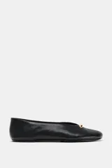 Bailarina plana de piel negra con acabado suave, escote en pico y detalle metálico dorado en la parte frontal. Presenta pespuntes decorativos y punta redonda.