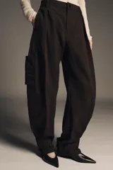 Pantalón cargo de algodón color marrón oscuro, tiro medio, con pliegues frontales, bolsillos delanteros ocultos y bolsillos de plastrón con solapa y botones a presión en las piernas. El bajo del pantalón tiene una trabilla con botón y se muestra doblado hacia afuera.