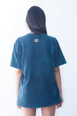 Remera de corte oversize con efecto de lavado ácido en color azul petróleo. Presenta un estampado gráfico frontal en color amarillo con tipografía moderna y la inscripción 'Montevideo, UY'.