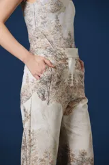 Pantalón de corte recto y tiro alto, de color beige con un estampado paisajístico de árboles y naturaleza en tonos marrones y grises. Cuenta con trabillas en la cintura y cierre frontal oculto.