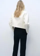 Chaqueta de corte recto y diseño estructurado en color crudo, con cuello redondo y manga larga. Presenta una abertura frontal sin cierre, decorada con ojales metálicos plateados a lo largo de los bordes. Confeccionada en tejido mezcla de algodón con forro interior.