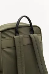 Mochila urbana de nylon en color verde militar, con detalles en cuero sintético negro. Cuenta con compartimento principal con cierre, bolsillo frontal con cierre, bolsillos laterales abiertos y asa superior. Incluye un colgante decorativo de tela y correas ajustables acolchadas.