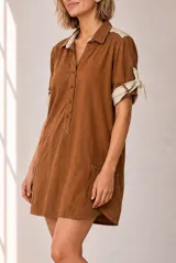 Vestido corto de lino color beige, de corte recto y holgado, con cuello polo y cierre frontal parcial con botones. Las mangas cortas cuentan con detalles de tiras para anudar en los puños.