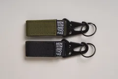 Llavero de cinta de poliéster color verde oliva con mosquetón y aro metálicos negros. Incluye etiqueta con el logo de la marca.