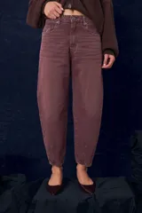 Jean de corte slouchy con tiro medio, confeccionado en denim de color bordó con efecto lavado. Presenta cierre frontal con botón y cremallera, bolsillos laterales y traseros, y un diseño de pierna relajada que se afina hacia el bajo con costuras verticales en los tobillos.