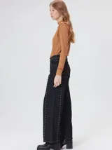 Jeans de corte ancho (wide leg) y tiro medio, confeccionados en denim de algodón negro con costuras contrastantes en color ocre. Presenta múltiples botones metálicos frontales y tachas metálicas aplicadas a lo largo de los laterales y la pretina.