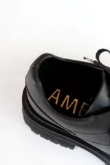 Zapatos de cuero negro con cordones, diseño robusto y suela track dentada. Presentan un detalle metálico en el centro de los cordones.