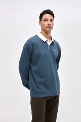 Remera polo de manga larga en color azul, confeccionada en algodón pesado con textura suave. Presenta cuello camisero en color crema, cierre frontal con botones y puños en las mangas. Incluye un pequeño bordado en el pecho.