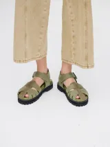 Sandalias romanas de cuero color verde militar, con diseño de tiras anchas entrelazadas, tira ajustable en el tobillo con hebilla metálica y suela dentada de goma negra.
