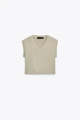 Jersey de punto color beige claro, con cuello en V y manga larga. Presenta detalles de botones decorativos en los hombros y en el ruedo inferior, sugiriendo un diseño desmontable o con aberturas ajustables.