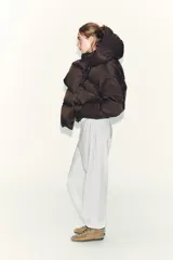 Campera puffer corta de color marrón oscuro, con diseño acolchado, cuello alto y capucha integrada. Presenta cierre frontal con botones magnéticos y mangas largas con puños elásticos.