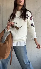 Sweater de punto color natural con cuello redondo y diseño de flores bordadas en las mangas. Presenta puños y ruedo acanalados.