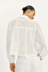 Blusa corta de color blanco con diseño de bordados perforados, cuello solapa, cierre frontal con botones y mangas largas abullonadas.