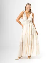Vestido largo color crudo, confeccionado en mezcla de tencel y lino, con escote en V pronunciado, breteles anchos terminados en lazos en los hombros, y falda larga de varios niveles con volados amplios y fluidos.