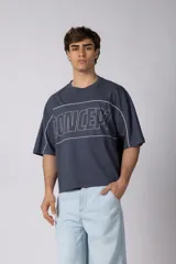 Remera gris oscuro de corte boxy, con mangas cortas y cuello redondo. Cuenta con un diseño gráfico frontal con líneas blancas y la palabra 'CONCEPT' estampada.