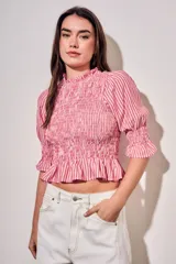 Blusa de algodón con diseño a rayas verticales en tonos rosa y blanco. Presenta un cuerpo con fruncido elástico (smock), cuello alto con volados y mangas tres cuartos abullonadas con terminación en volado.