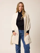 Poncho abierto de tejido de punto en color gris claro con bordes y detalles de bolsillos en gris oscuro. Presenta un diseño holgado y versátil con placa metálica decorativa en uno de los bolsillos frontales.