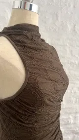 Vestido corto marrón con textura arrugada, cuello alto asimétrico y una manga larga. Presenta un corte en la cintura.